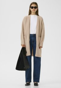 Moteris, dėvinti juodus akinius nuo saulės, smėlio spalvos atvirą kardiganą, baltą marškinėlį, mėlynus džinsus, juodus butus, laiko didelę juodą rankinę ant balto fono.