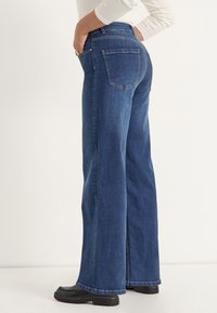 Donkerblauwe wijde jeans van denim. Kenmerken zijn onder andere twee achterzakken en een subtiele vervaagde textuur. Draagt met zwarte platform schoenen.