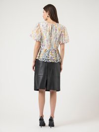 Vrouw die een bloes met pofmouwen en bloemenprint draagt, met een gelaagde zoom en een zwarte lederen rok tot op de knieën, staande met de rug naar de camera en zwarte hakken.