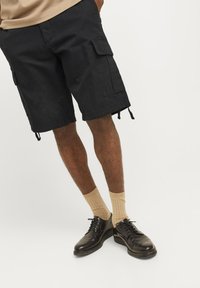 Sorte cargo shorts med flere sidelommer, snøre i bunden og afslappet pasform, parret med beige ribbede sokker og sorte sko.