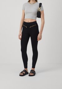 Mustat legginsit, joissa on kaksi edessä vetoketjullista taskua, yhdistettyinä istuvaan harmaaseen lyhytpaidan ja mustiin remmisandaaleihin. Sileä tekstuuri, slim fit.