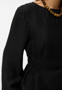 Melna struktēta blūze ar garām piedurknēm, ar savāktu vidukli un apaļu kakla izgriezumu, kombinēta ar lielu, apaļu krellēta kaklarotu.