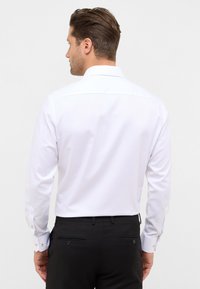 Man die met zijn rug naar de camera staat, gekleed in een wit overhemd met lange mouwen en zwarte broek, tegen een eenvoudige witte achtergrond.