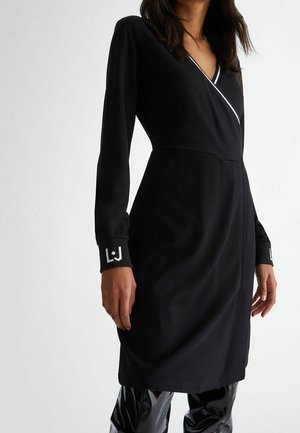 Femme portant une robe portefeuille noire avec des bordures blanches et un logo sur les poignets, associée à des bottes noires brillantes montant jusqu'aux genoux, se tenant devant un fond uni.