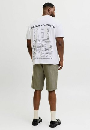 Homme de dos portant un T-shirt blanc avec un dessin au trait noir d'un café et un texte, un short olive, des chaussettes blanches et des chaussures noires.