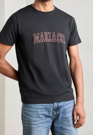 Mand iført sort t-shirt med korte ærmer med lyserød "MAKIA CO." tekst og lyseblå cowboybukser, står med den ene hånd bag ryggen.