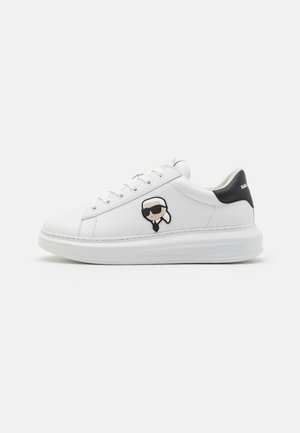 Witte lage sneaker met zwarte hieltab, veters en een cartoon gezichtslogo met zonnebril aan de zijkant.
