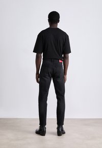 T-shirt noir vu de l'arrière, associé à un jean noir. Le jean présente une étiquette rouge sur la poche arrière, complétant des bottines noires.