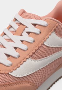 Κοντινό πλάνο ροδακινί αθλητικού παπουτσιού με λευκό δίχτυ, λευκά κορδόνια και κυματιστό διακοσμητικό κατά μήκος της περιοχής των κορδονιών.