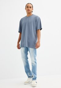 Camiseta gris de gran tamaño con mangas cortas, combinada con jeans azul claro y zapatillas crema. Diseño simple y casual con un ajuste relajado.
