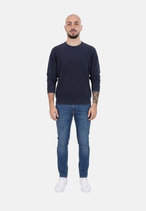 Uomo in piedi rivolto verso avanti, indossa un maglione blu navy, jeans blu, scarpe da ginnastica bianche, con un tatuaggio visibile sull'avambraccio sinistro.