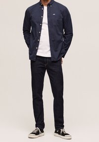 Camisa navy de botones sobre una camiseta blanca, jeans oscuros y zapatillas negras. Presenta un bolsillo en el pecho y está hecha de tela de algodón.