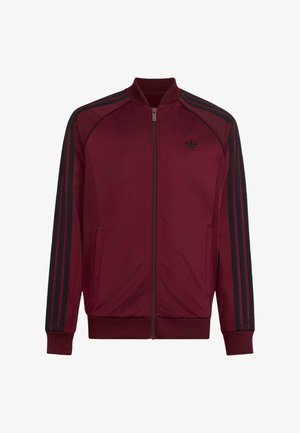 Chaqueta deportiva Adidas color granate con cremallera, tres franjas negras en las mangas y un pequeño logotipo negro de Adidas en el pecho izquierdo.