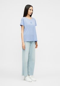 Γυναίκα που στέκεται φορώντας ανοιχτό μπλε κοντομάνικη μπλούζα, τζιν παντελόνι με φαρδιά μπατζάκια σε ανοιχτή απόχρωση και λευκά αθλητικά παπούτσια, πάνω σε ένα απλό φόντο.