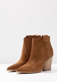 Bottines en daim marron avec bouts pointus, motifs de couture décoratifs et un talon empilé. Comprend des languettes pour un enfilage facile.
