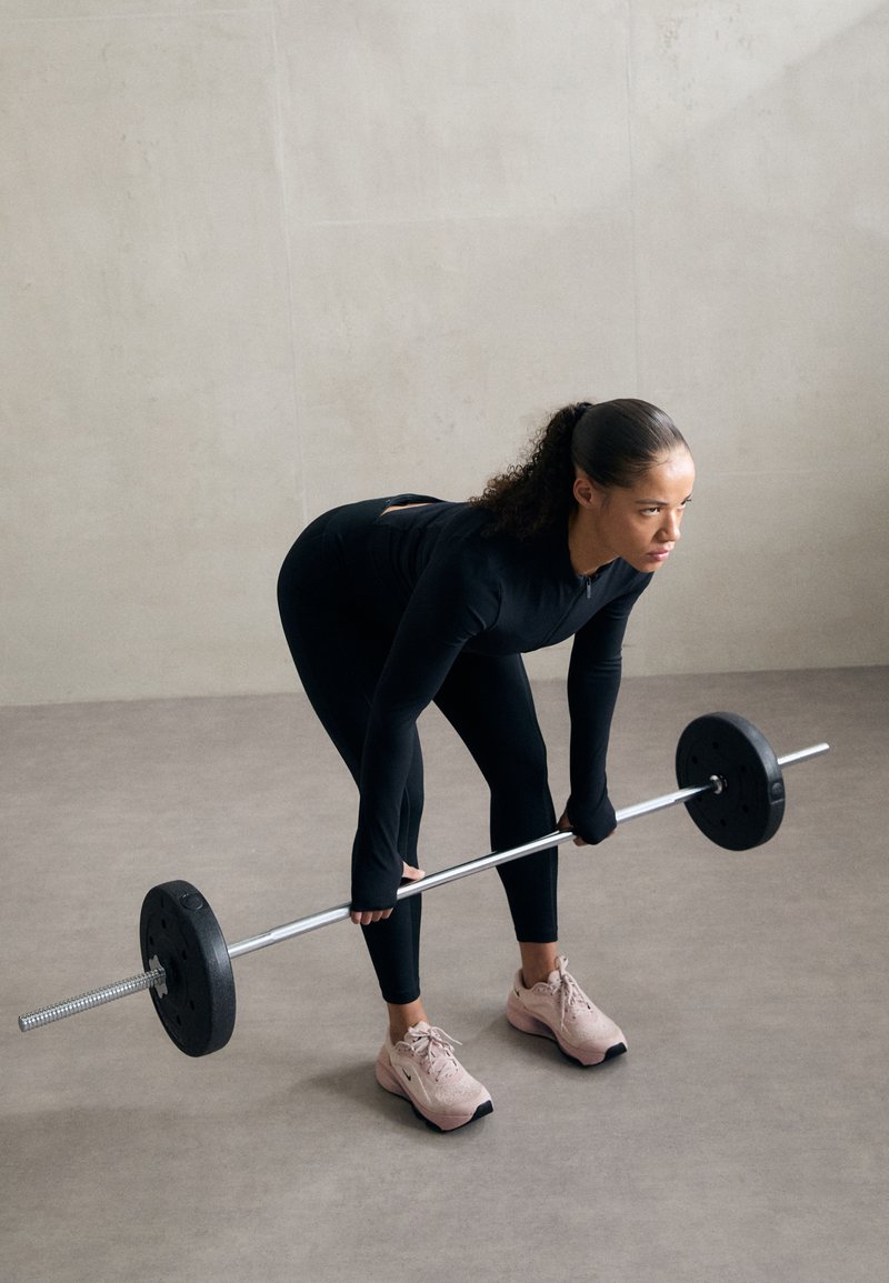 Schwarzes Sportoutfit mit langen Ärmeln, figurbetontem Design und einem Ausschnitt-Detail. Das Subjekt hebt eine silberne Hantel mit schwarzen Gewichten. Rosa Turnschuhe.