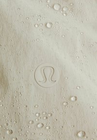 Atsparus vandeniui audinys šviesiame beige su tekstūruota paviršiumi ir daugybe vandens lašelių. Apačioje dešinėje yra skaidrus logotipas.
