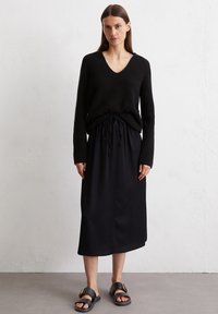 Schwarzer, gerippter V-Ausschnitt-Pullover, kombiniert mit einem schwarzen Rock mit Kordelzug in der Taille. Dazu schwarze Sandalen mit einem goldenen Schnallendetail.