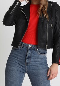 Giacca in pelle nera da motociclista con cerniere argentate, indossata su un maglione a coste rosso brillante. Jeans a vita alta blu chiaro con cuciture visibili.