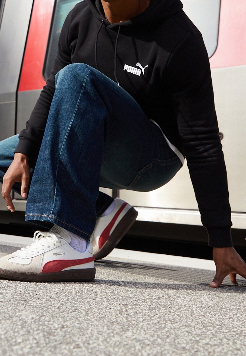 Bílé tenisky s červenými akcenty, s texturovaným povrchem a gumovou podrážkou, kombinované s tmavě modrými džíny a černou mikinou.