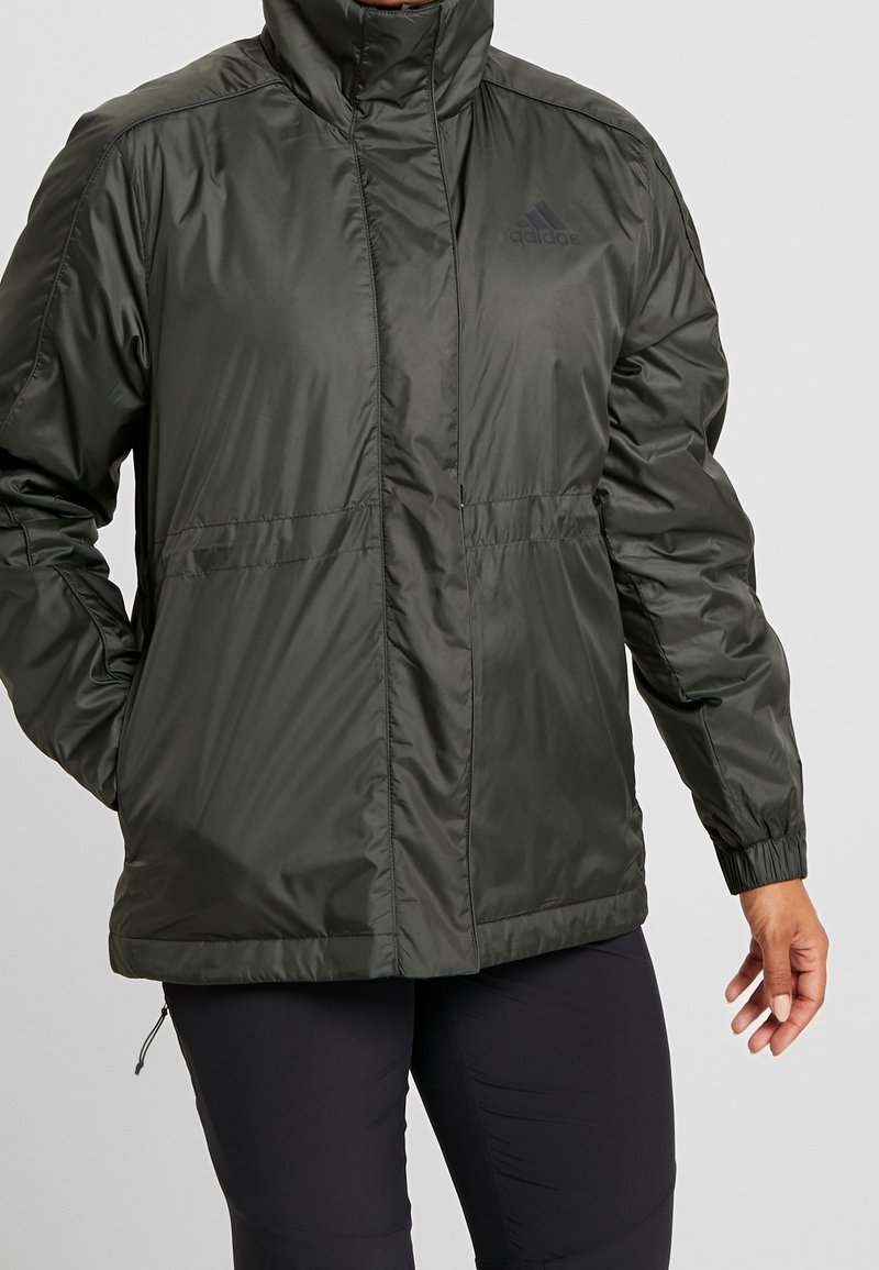 Veste légère noire Adidas avec col montant, poignets élastiques, mains dans les poches, portée avec un pantalon noir.