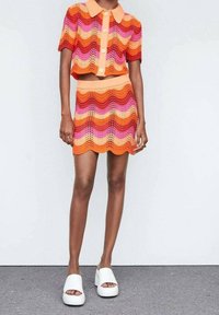 Modèle portant un ensemble en deux pièces coloré, crocheté en orange, rose et rouge, avec des bords festonnés et des sandales à plateforme blanches.