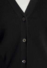 Sort ribbet cardigan med tre sorte knapper over en hvid ribbet top, der viser bryst- og øvre torsoområde.