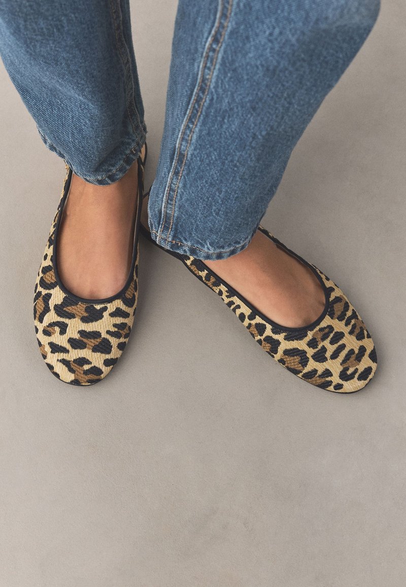 Füße, die Leopardenmuster-Ballerinas tragen, kombiniert mit blauen Jeans auf einem hellgrauen Boden.