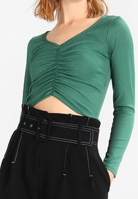 Green long-sleeve top with a V-neck, featuring gathered texture at the center. Paired with high-waisted black pants and a wide belt.