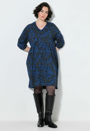 Donna con capelli corti e scuri che indossa un vestito nero e blu con motivo fino al ginocchio, collant neri e stivali neri al ginocchio, in piedi con le mani dietro la schiena.