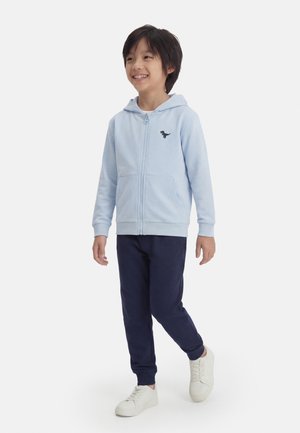 Lachende jongen loopt, draagt een lichtblauwe zip-hoodie met dinosauruslogo, donkerblauwe broek en witte sneakers op een effen achtergrond.