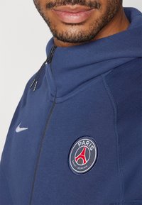 Sweat-shirt bleu marine avec un col zippé, arborant un logo Nike et un emblème rond du Paris Saint-Germain sur la poitrine ; texture lisse.