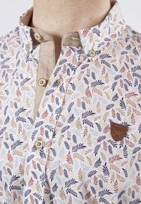 Chemise blanche ornée d'un motif de feuilles dans des tons de bleu, d'orange et de vert. Comprend des détails beiges, une fermeture à boutons et une étiquette de logo marron.