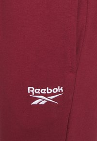 Rubinröda Reebok-joggingbyxor i ett mjukt tyg, med en vit broderad logo och en figurnära design med sidostygnsdetaljer.