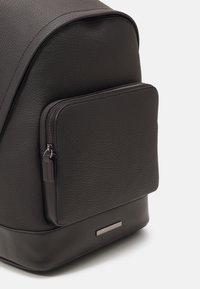 Zaino nero testurizzato con una base liscia e una tasca frontale con zip. Presenta dettagli metallici e una forma rettangolare.