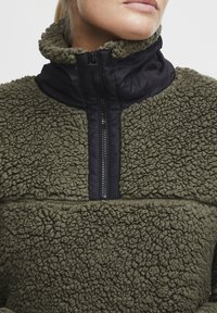 Olijfgroen fleece pullover met een zwarte nylon ritssluiting op de kraag. Gevoerde structuur, half-rits ontwerp en contrasterende materialen accenten.