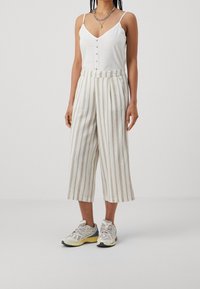 Camisola bianca leggera con bottoni e spalline sottili, abbinata a pantaloni a gamba larga a strisce beige e sneakers sportive multicolori.