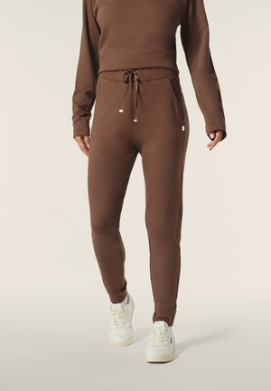 Pantalon de survêtement marron en tissu doux, doté d'une taille avec cordon de serrage, de poches latérales et d'une coupe fuselée, associé à des baskets blanches.