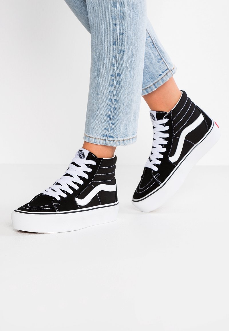 Baskets montantes noires avec lacets blancs et bandes latérales portées avec un jean bleu clair retroussé sur fond blanc.