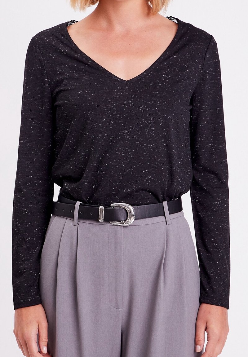 Femme portant un haut noir à manches longues avec col en V, rentré dans un pantalon gris taille haute avec une ceinture noire et une boucle argentée.