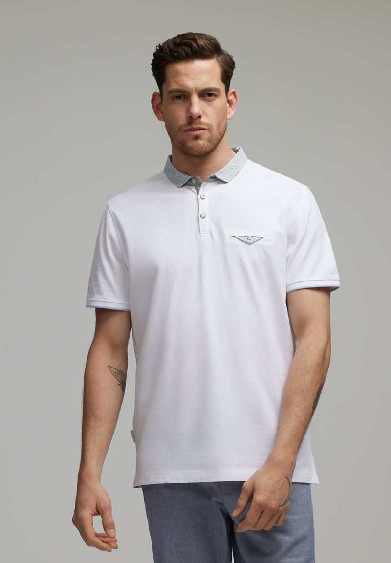 Biała koszulka polo wykonana z bawełny, z wzorzystym niebieskim kołnierzykiem, z dwoma guzikami oraz małą kieszonką na piersi. Krótkie rękawy.