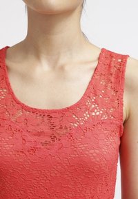 Haut en dentelle rouge avec un col rond et sans manches. La dentelle présente des motifs floraux et une finition texturée.