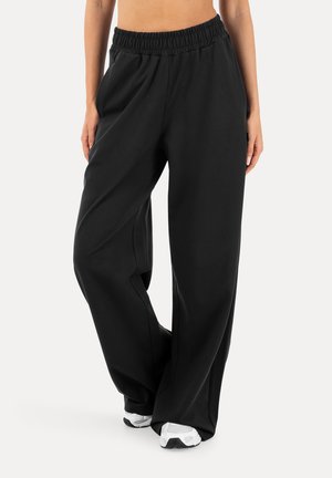 Osoba ubrana w luźne, czarne szerokie spodnie z elastycznym paskiem i białe trampki, stojąca na jednolitym tle.