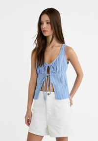 Světle modrý bezrukávový top s bílými pruhy, s předními vázacími detaily a výstřihem ve tvaru V. Vyznačuje se hladkou strukturou a uvolněným střihem. Kombinováno s bílými šortkami.