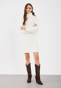 Witte kabelgebreide sweaterjurk met hoge halslijn, geribbelde zoom en lange mouwen, gecombineerd met donkerbruine cowboylaarzen met patroon.