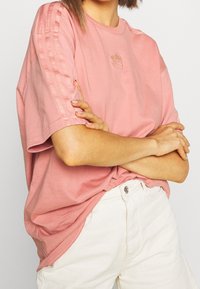 Rosa överstor t-shirt med korta ärmar, har tre kontrasterande ränder på varje axel och en liten logotyp på bröstet. Bärs med vita byxor.