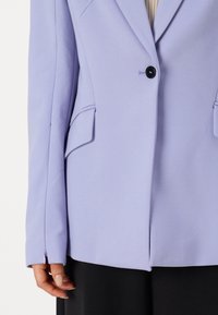 Donna che indossa un blazer violaceo chiaro con un bottone nero singolo e tasche a patta, abbinato a pantaloni neri, in piedi con la mano rilassata.