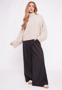 Kobieta w beżowym swetrze z golfem, szerokich czarnych spodniach i beżowo-białych sneakersach stoi na tle jasnego, jednolitego tła.