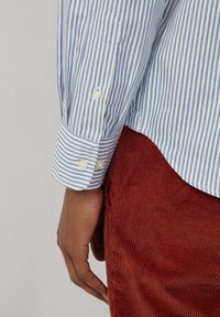 Camisa a rayas azul y blanca con puños abotonados, combinada con pantalones de pana color óxido. La camisa presenta una textura suave y un corte ajustado.