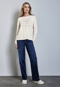 Langärmliges gestreiftes Creme-Hemd mit einem grafischen Druck, kombiniert mit dunkelblauen Jeans und hellen Sneakers auf einer flachen Oberfläche.